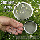 Two metal straining sieves held by a hand on grass with flowers